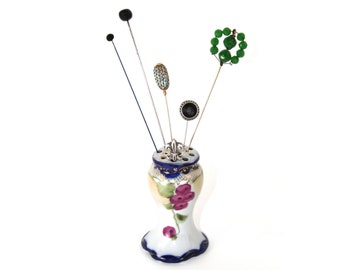 May include: A ceramic pin cushion holder with a white base, painted purple floral motifs, and a blue rim, topped with a silver fleur-de-lis. Four decorative hat pins are inserted into the top, featuring black spheres, a textured blue bead, a black disc, and a green beaded circle.