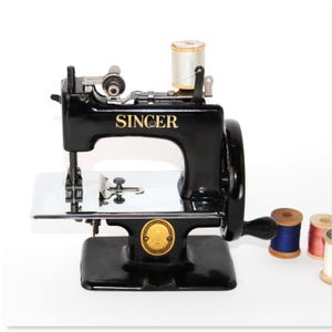 May include: A vintage black Singer sewing machine with gold lettering. The machine has a silver needle plate and a decorative emblem. Several spools of thread in various colours are placed to the right of the machine.