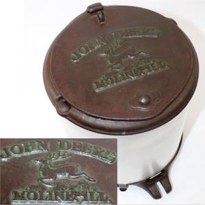 Puede incluir: Un desgranador de maíz vintage de hierro fundido John Deere con acabado de esmalte blanco y tres patas. La tapa tiene un logotipo en relieve verde y marrón con el texto "John Deere" y "Moline, Ill."
