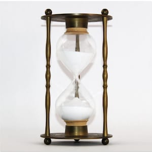 Könnte beinhalten: Eine Sanduhr im antiken Stil mit einer durchsichtigen Glaskugel, die mit weißem Sand gefüllt ist und sich in einem messingfarbenen Metallrahmen befindet. Die Sanduhr hat ein klassisches Design mit einem runden Ober- und Unterteil, verbunden durch geschwungene Stützen. Der Sand fließt.