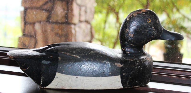 May include: A black and white wooden duck decoy with a white band around the middle. The decoy has a black head and a long, thin beak.