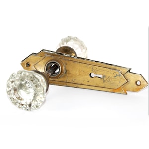 May include: A door handle set featuring two clear, faceted glass knobs attached to a rectangular, weathered brass backplate. The backplate has a central keyhole, a circular spindle opening, and pointed ends with screw holes for mounting.