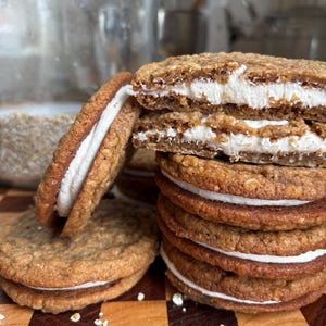 Puede incluir: Una pila de galletas de avena con crema que consta de dos galletas de avena doradas y texturizadas unidas por una capa gruesa de relleno de crema blanca. Una galleta está partida por la mitad, revelando su textura interior suave.