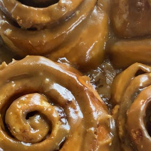 May include: A close-up view of several spiral-shaped cinnamon rolls covered in a thick, glossy, light brown caramel glaze. The pastry dough is visible in the center of each roll, showing a golden-brown texture beneath the smooth, shiny coating.