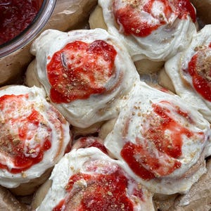 May include: A batch of baked sweet rolls arranged in a parchment-lined pan, topped with a thick layer of white cream cheese frosting, a drizzle of bright red strawberry fruit compote, and a light dusting of golden brown cookie crumbs.