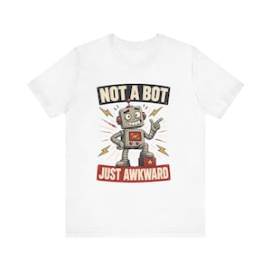 Könnte beinhalten: Ein weißes kurzärmeliges T-Shirt mit einer Grafik eines grauen Roboters, der auf einem roten Block steht. Der Roboter zeigt mit einer Hand. Über dem Roboter steht der Text NOT A BOT und unter dem Roboter der Text JUST AWKWARD in roten Buchstaben.