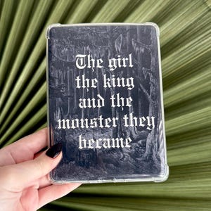 Peut inclure: Une coque de protection rectangulaire transparente pour appareil électronique avec une illustration de forêt sombre et détaillée et le texte gothique blanc : The girl the king and the monster they became, imprimé au dos de la coque.