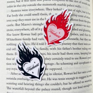 May include: Two heart-shaped stickers with flame designs. One sticker features a red flame surrounding a light pink heart with the text Fireheart in cursive. The other sticker features a black flame surrounding a white heart with the text Fireheart.