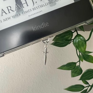 May include: A silver-colored metal sword charm attached to the bottom edge of a Kindle e-reader. The screen displays text including the name Sarah J. Maas and the word Bloomsbury. The device is encased in a clear protective cover.