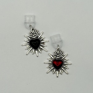 May include: A pair of silver-toned metal earrings featuring a sacred heart design with radiating spikes. One heart is filled with black enamel, and the other is filled with red enamel. Each earring includes a clear plastic earring back attachment.