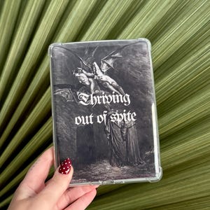 May include: A clear rectangular phone case featuring a black and white gothic illustration of winged figures and cloaked individuals. The text "Thriving out of spite" is printed in a white gothic font across the center of the design.