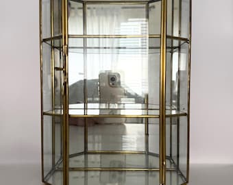 May include: A tall, hexagonal display case featuring clear glass panels framed in brass. The structure includes two internal glass shelves and a hinged door with a latch on the left side. The base and top are also made of glass.