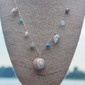 May include: A silver chain necklace featuring a central round peach-colored shell pendant, accented with smaller scattered seashells, smooth round beads in shades of turquoise, pale green, and soft pink, all suspended along the delicate metal links.