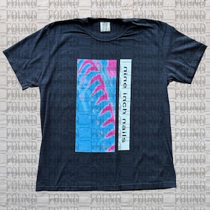 May include: A black short-sleeve t-shirt featuring a rectangular graphic on the chest. The graphic displays a blue background with a series of pink, curved, claw-like shapes and a vertical white bar containing the black text nine inch nails.