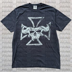 May include: A black short-sleeve t-shirt featuring a white graphic print of a stylized skull with large curved horns centered over a cross shape. The shirt has a crew neckline and a standard fit, with a visible brand tag at the inner collar.