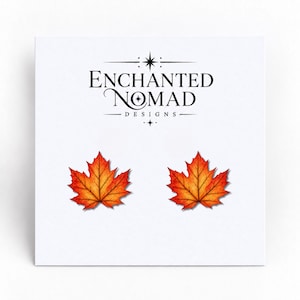 May include: A pair of stud earrings shaped like maple leaves with vibrant orange, red, and yellow gradients. The earrings are displayed on a white card featuring the black text Enchanted Nomad Designs with a star icon above and below the brand name.