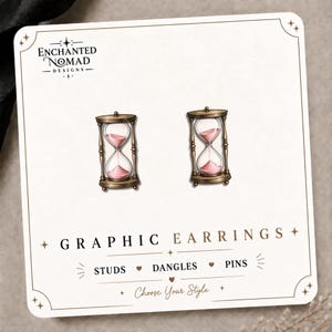 May include: A pair of stud earrings featuring an illustration of an hourglass with a bronze frame and pink sand. The earrings are displayed on a white card labeled Enchanted Nomad Designs, Graphic Earrings, Studs, Dangles, Pins, Choose Your Style.