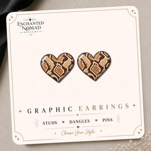 May include: A pair of heart-shaped stud earrings featuring a brown and tan snakeskin pattern. The earrings are displayed on a white card labeled Enchanted Nomad Designs with the text Graphic Earrings, Studs, Dangles, Pins, and Choose Your Style.