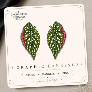 May include: A pair of leaf-shaped earrings featuring a dark green surface with white spots and a pink underside, displayed on a white card labeled Enchanted Nomad Designs Graphic Earrings with options for studs, dangles, and pins.