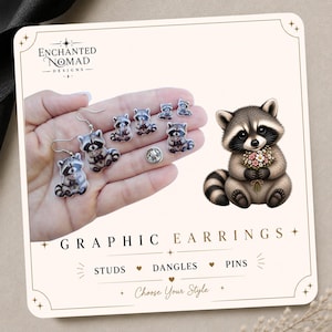 May include: A display card titled Enchanted Nomad Designs featuring graphic earrings, studs, and pins. The card shows a raccoon illustration holding flowers and various raccoon-themed jewelry pieces, including dangle earrings and small stud pins.
