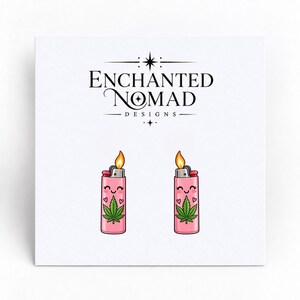 May include: A pair of stud earrings featuring a pink lighter design with a green cannabis leaf, a smiling face, and small hearts. Above the earrings, the text reads ENCHANTED NOMAD DESIGNS with a star icon centered between the words.