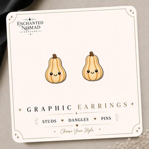 May include: A pair of light orange pumpkin stud earrings with a smiling face graphic on a white display card. The card features the text Enchanted Nomad Designs, Graphic Earrings, and lists options for Studs, Dangles, and Pins with Choose Your Style.