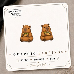 May include: A pair of stud earrings featuring a brown capybara holding a taco, displayed on a white card with the text Enchanted Nomad Designs, Graphic Earrings, Studs, Dangles, Pins, and Choose Your Style printed in black and gold lettering.