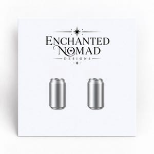 May include: A pair of silver-colored metal soda can-shaped stud earrings displayed on a white card featuring the black text Enchanted Nomad Designs with a star icon above and below the brand name on a plain white background.
