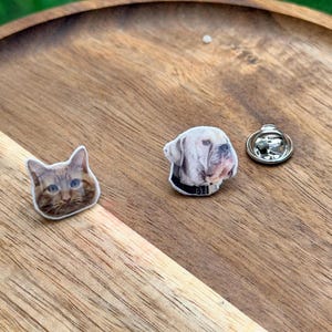 May include: Two small lapel pins featuring the head of an orange tabby cat and the head of a light-colored dog wearing a black collar. The pins are flat with printed images and include a silver-toned metal clutch backing for attachment.