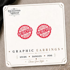 May include: A pair of circular stud earrings featuring a red graphic stamp design with the word REJECTED printed in a distressed font. The earrings are displayed on a white card labeled Enchanted Nomad Designs with the text Graphic Earrings.