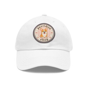 May include: A white baseball cap featuring a circular patch on the front. The patch has a light pink background with a cartoon illustration of a tan and white Corgi dog, surrounded by the text "perridise shop" and small heart and paw print icons.