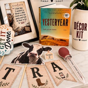 May include: A Yesteryear decor kit featuring a framed quote, a book titled Yesteryear by Caro Claire Burke, a cow cutout, a Sassafras 2 tag, a Get It Done sign, lettered pennants spelling TRA, and a baster labeled Emotional Support Baster.