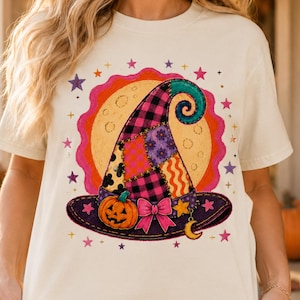 May include: A cream-coloured t-shirt featuring a graphic of a patchwork witch hat in shades of pink, purple, orange, and teal. The hat is decorated with a jack-o'-lantern, a pink bow, and a gold crescent moon charm against a large yellow moon.