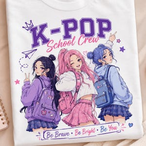 Può includere: Una t-shirt bianca con una grafica di tre personaggi in stile anime con zaini e il testo K-POP School Crew. Il design include colori viola, rosa e blu, stelle, una corona e la frase Be Brave, Be Bright, Be You.