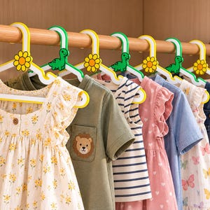May include: A row of white plastic clothes hangers with colorful decorative accents hanging on a wooden rod. The hangers feature alternating designs of yellow flowers and green dinosaurs at the hook, supporting various children's garments.
