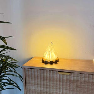 May include: A decorative lamp shaped like a campfire with a translucent white flame element resting on a base of small, dark brown cylindrical logs. The lamp emits a warm, soft yellow glow, creating a cozy light effect in the surrounding area.