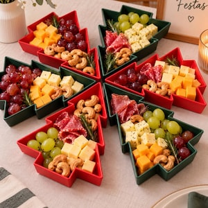 May include: Six Christmas tree-shaped serving dishes in red and dark green, arranged in a circular pattern. Each dish is filled with a variety of snacks including cubed yellow and white cheeses, red and green grapes, salami slices, and roasted cashews.