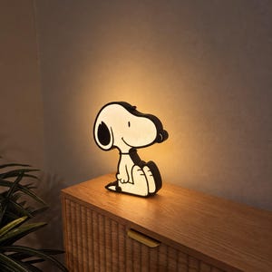 May include: A Snoopy-shaped lamp with a black outline and a warm, glowing white center. The lamp features the iconic cartoon dog sitting in profile, emitting a soft light that illuminates the surrounding area with a gentle, golden-toned glow.