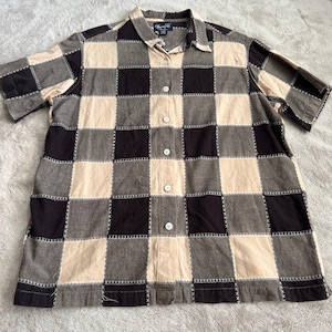 May include: A short-sleeved button-down shirt featuring a large checkered pattern in alternating squares of dark charcoal gray and light beige. The seams between each square are highlighted with a decorative white cross-stitch embroidery detail.
