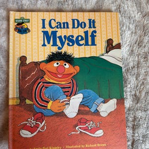 May include: A hardcover children's book titled I Can Do It Myself, featuring an illustration of Ernie from Sesame Street sitting on a floor, putting on white socks, with red sneakers nearby and a bed in the background.