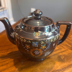 May include: A dark brown ceramic teapot with a glossy finish, featuring a lid with a knob handle. The body is decorated with a horizontal band of gold-toned vertical lines and small, colourful floral patterns in blue, orange, and yellow.
