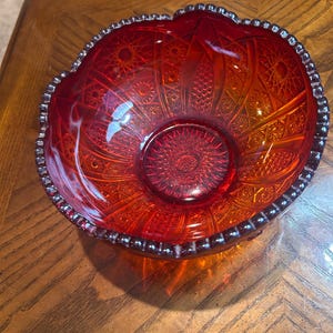 Puede incluir: Un cuenco de cristal rojo intenso con un complejo patrón prensado de estrellas, sombreado cruzado en forma de diamante y paneles verticales. El cuenco tiene un borde festoneado con cuentas y una base circular con un diseño de estrella central translúcido.