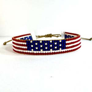 May include: A woven beaded bracelet featuring a pattern of horizontal red and white stripes with a rectangular blue section containing small white beads. The bracelet has adjustable tan cord ties on both ends to secure it around the wrist.