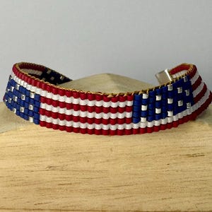 May include: A woven beaded bracelet featuring a repeating pattern of the American flag with horizontal red and white stripes and a blue section with silver beads representing stars. The bracelet has a metallic clasp and a flexible, flat construction.