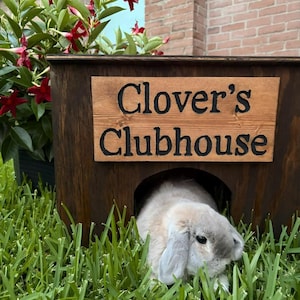 May include: A dark brown wooden pet house with a rectangular sign featuring the text Clover's Clubhouse in black lettering. A light-colored lop-eared rabbit is positioned inside the arched entrance of the structure, surrounded by green grass.