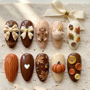 May include: A set of ten almond-shaped press-on nails featuring autumn-themed 3D designs including a gingerbread man, a pumpkin, a coffee mug, maple leaves, acorns, buttons, a cable knit texture, and bows, accented by a cream-coloured ribbon.