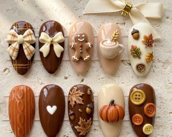 May include: A set of ten almond-shaped press-on nails featuring autumn-themed 3D designs including a gingerbread man, a pumpkin, a coffee mug, maple leaves, acorns, buttons, a cable knit texture, and bows, accented by a cream-colored ribbon.