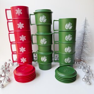 May include: A set of stackable plastic mugs in red and green. The red mugs feature white snowflake designs, while the green mugs display white bird motifs. Each mug includes a matching lid, with several lids stacked separately in front of the mugs.