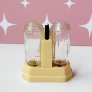 May include: A vintage salt and pepper shaker set featuring two clear, vertically ribbed glass containers held within a cream-colored plastic base. The base includes a central vertical handle with a black accent at the top, connecting the two shakers.
