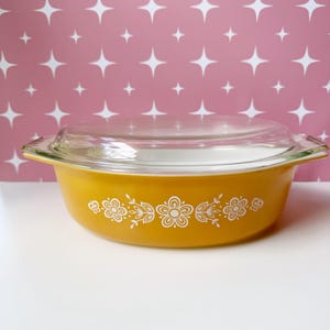 May include: A round, golden-yellow casserole dish with a clear glass lid. The exterior of the dish features a white floral pattern consisting of stylized flowers and leaves arranged in a horizontal band around the center of the vessel.